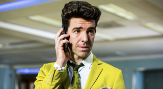 Paul Ready wears a mustard yellow suit and is on the phone in Utopia