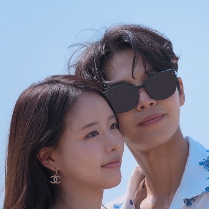 A woman (Park Hee-sun) wears Chanel logo earrings and a man (Lim Su-been) wears a white collared shirt with blue printed flowers and sunglasses. The couple poses for a selfie as he holds up a white instant camera, in a still from 'Single's Inferno' season 5. 