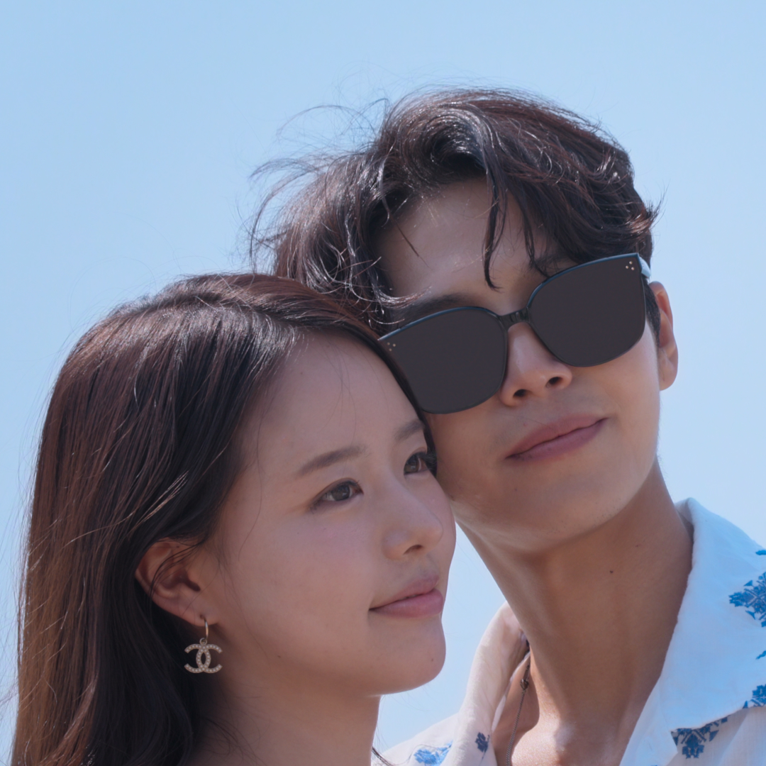 A woman (Park Hee-sun) wears Chanel logo earrings and a man (Lim Su-been) wears a white collared shirt with blue printed flowers and sunglasses. The couple poses for a selfie as he holds up a white instant camera, in a still from 'Single's Inferno' season 5. 