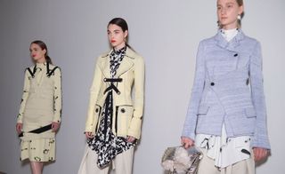 Three female models, one in a cream dress with black details, one in a black and white blouse with a cream jacket, and one in a pale blue coat