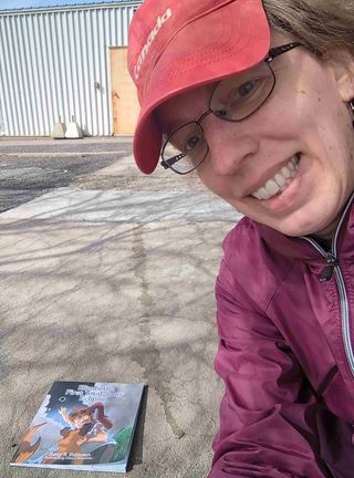 a selfie of a woman in a coat and hat. on the ground are shadows of tree branches and a children's book that says "Elizabeth's First Total Solar Eclipse" on the front