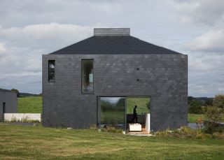 A square black timber clad home in a green field 