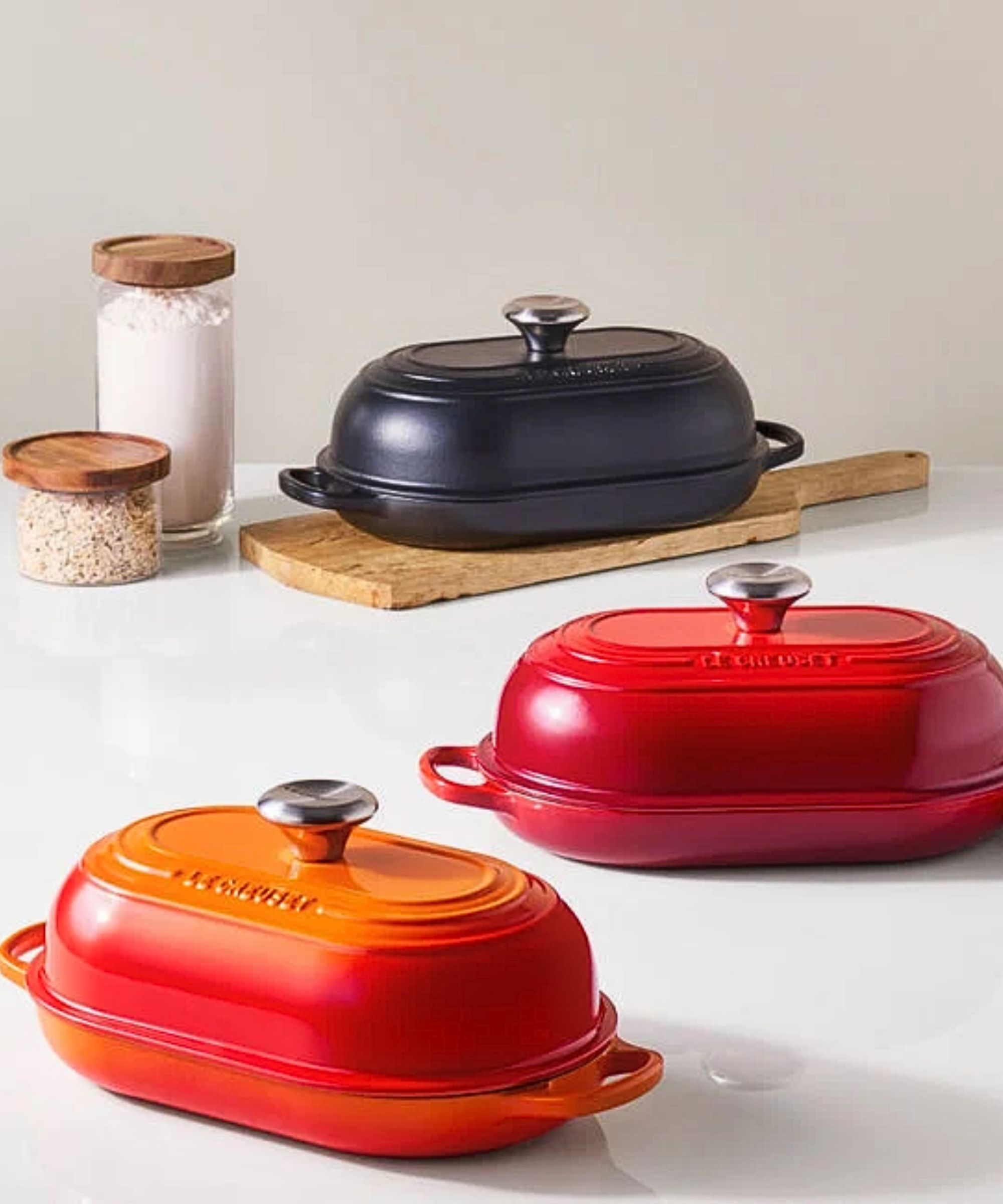 Three Le Creuset oval bread ovens, in orange, red, and navy blue on a white countertop.