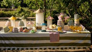 Image of a long outdoor table with a green and white striped tablecloth covered in different colorful lanterns, lamps, and lampshades.