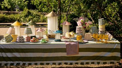 Image of a long outdoor table with a green and white striped tablecloth covered in different colorful lanterns, lamps, and lampshades.