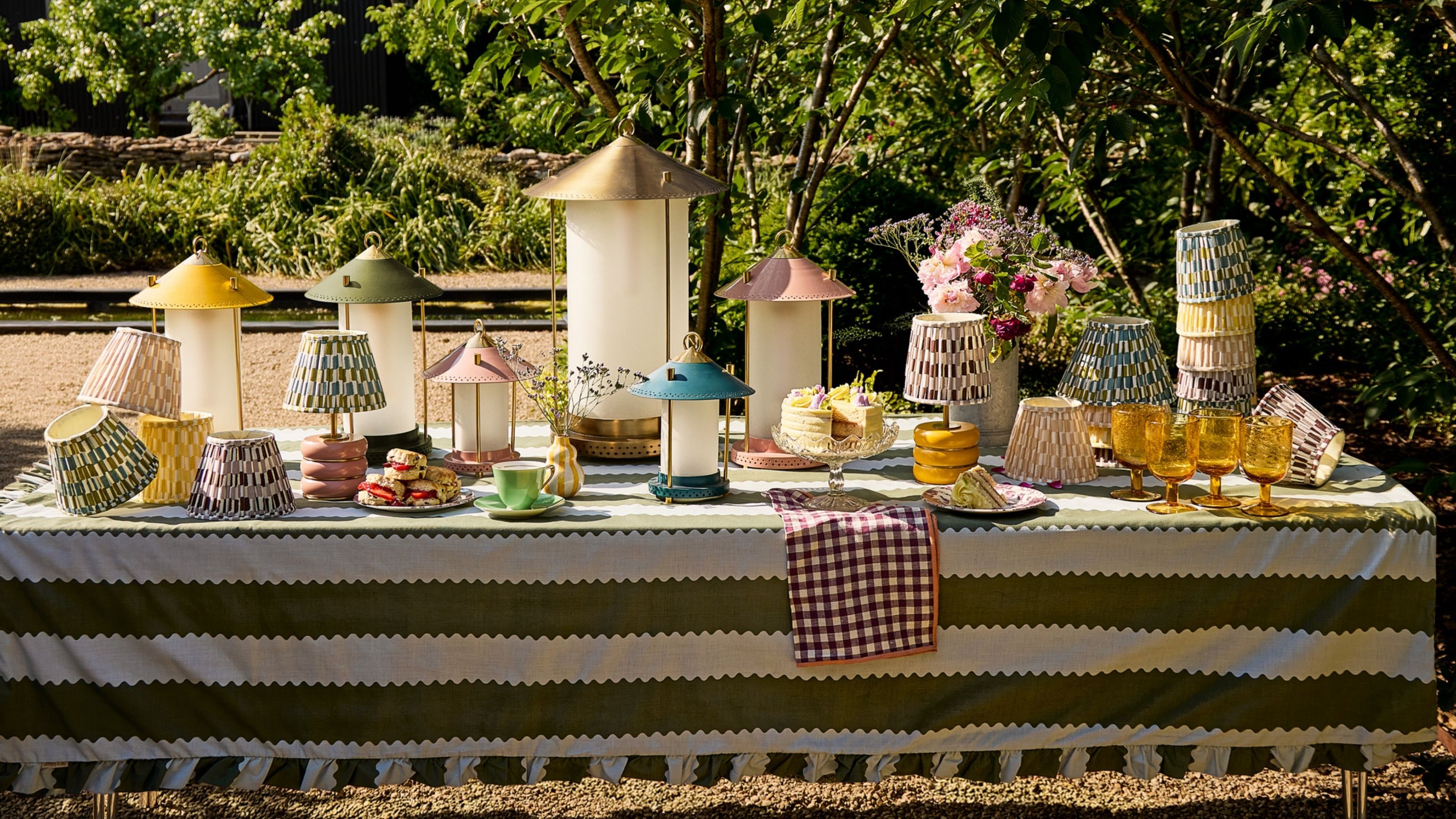 Image of a long outdoor table with a green and white striped tablecloth covered in different colorful lanterns, lamps, and lampshades.