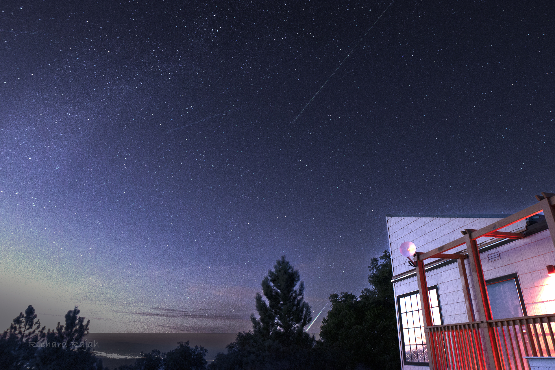 Perseid meteors dot the sky and a bright torch trails behind the trees in the lower part of the image.  A building is illuminated on the right of the image.