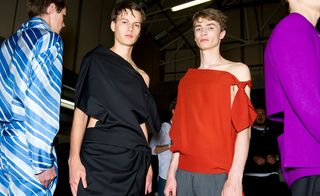 Four guys wearing the JW Anderson S/S 2015 collection. On the left the guy is wearing a blue and gray one piece. Next to him the guy is wearing a black off the shoulder draped garment. The guy next to him is wearing a red off the shoulder top with gray pants and on the right is a guy wearing a purple top.