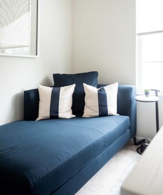 A white room with white walls with a white wall art print, a navy blue rectangular chaise longue with white and blue striped pillows, a window, and the corner of a desk