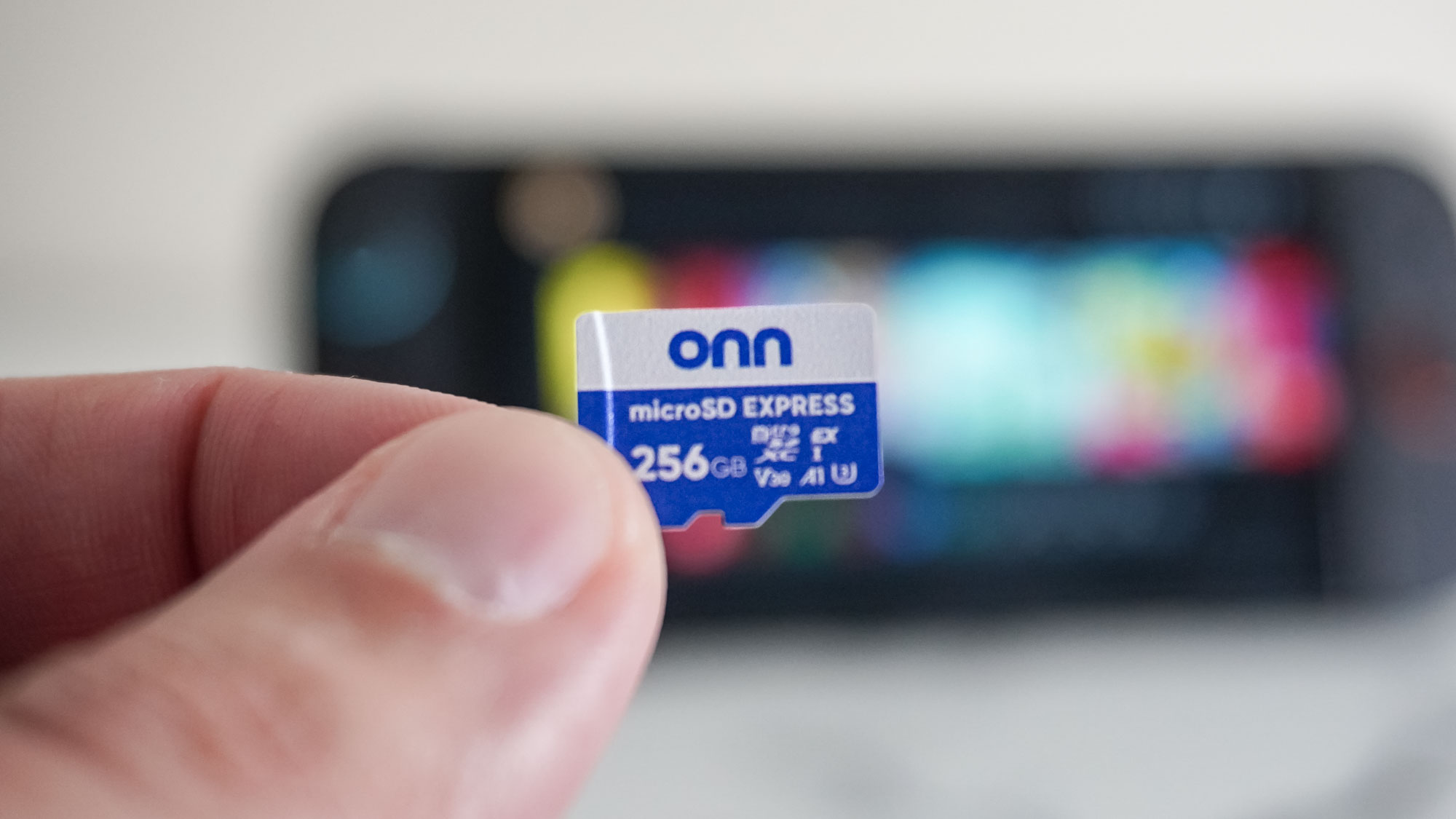 A person holding a microSD Express card in the foreground with a blurred Nintendo Switch 2 on a desk in the background