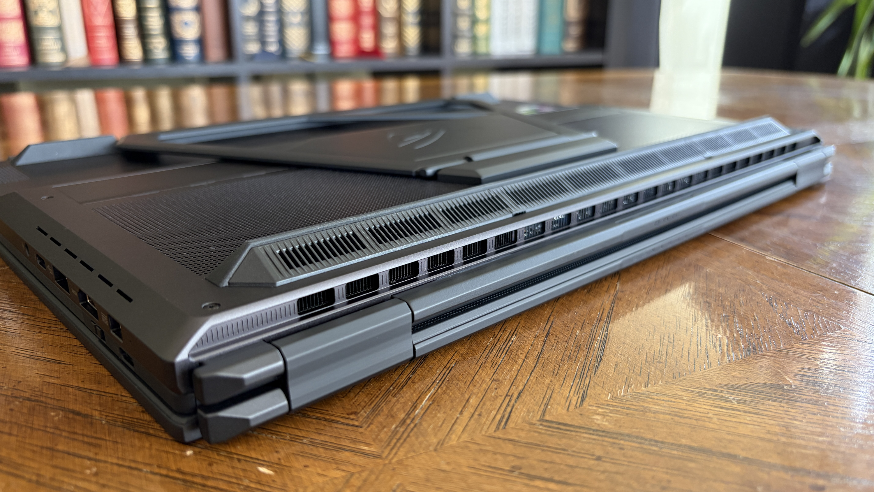 A sleek gaming laptop sits closed on a wooden table, highlighting its angular design and cooling vents. Blurred bookshelves are visible in the background.