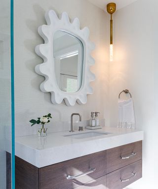 Relaxed bathroom space with large mirror in white with rounded, scalloped edging, brass and opal slender hanging pendant, dark wood vanity cabinet and sink with marble countertop