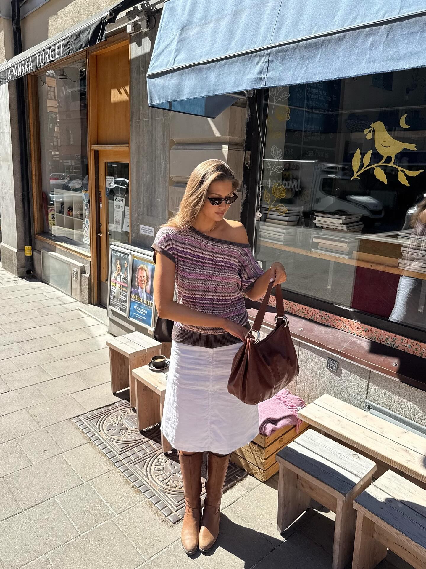 Fashion person Sofia wears a stripped of the shoulder tee, a white linen skirt and wears brown rider boots. She is stood infront of a book shop in a recent image from her Instagram.