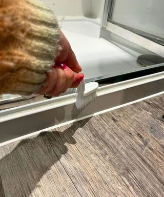 Hand holding scrubbing brush and cleaning a narrow gap between shower tray and chrome shower door frame