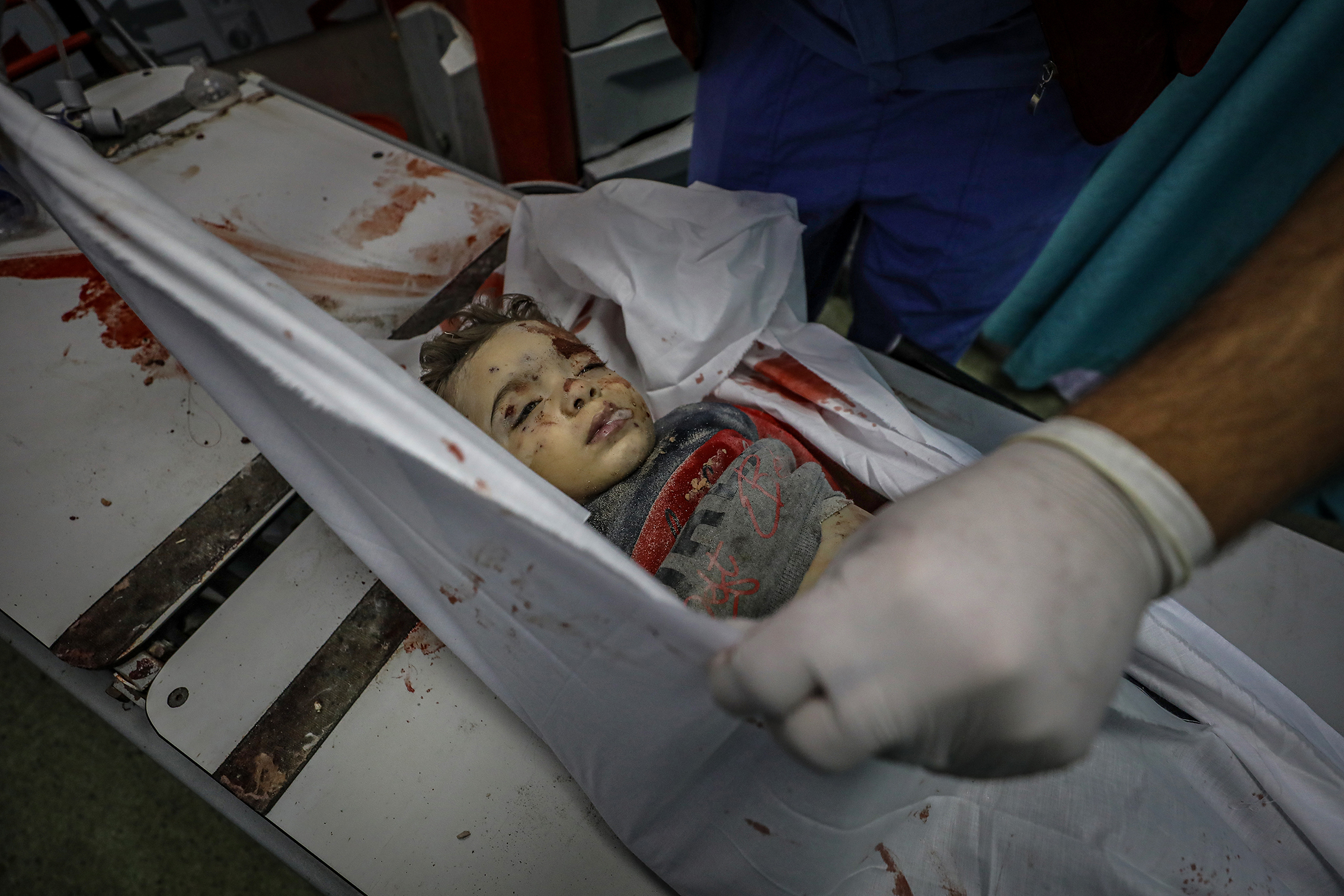 Image taken inside a Gaza hospital morgue following an airstrike, showing a dead boy with injuries, wrapped in a white cloth that gets handled by a medical person