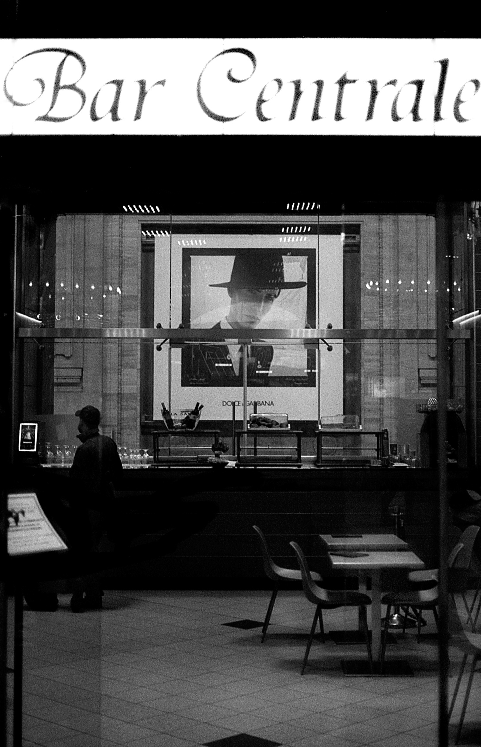 Large Dolce &amp;amp; Gabbana poster of a man in a wide-brimmed hat hangs behind the counter of a near-empty bar at night, beneath an illuminated "Bar Centrale" sign, as a lone customer stands at the counter.