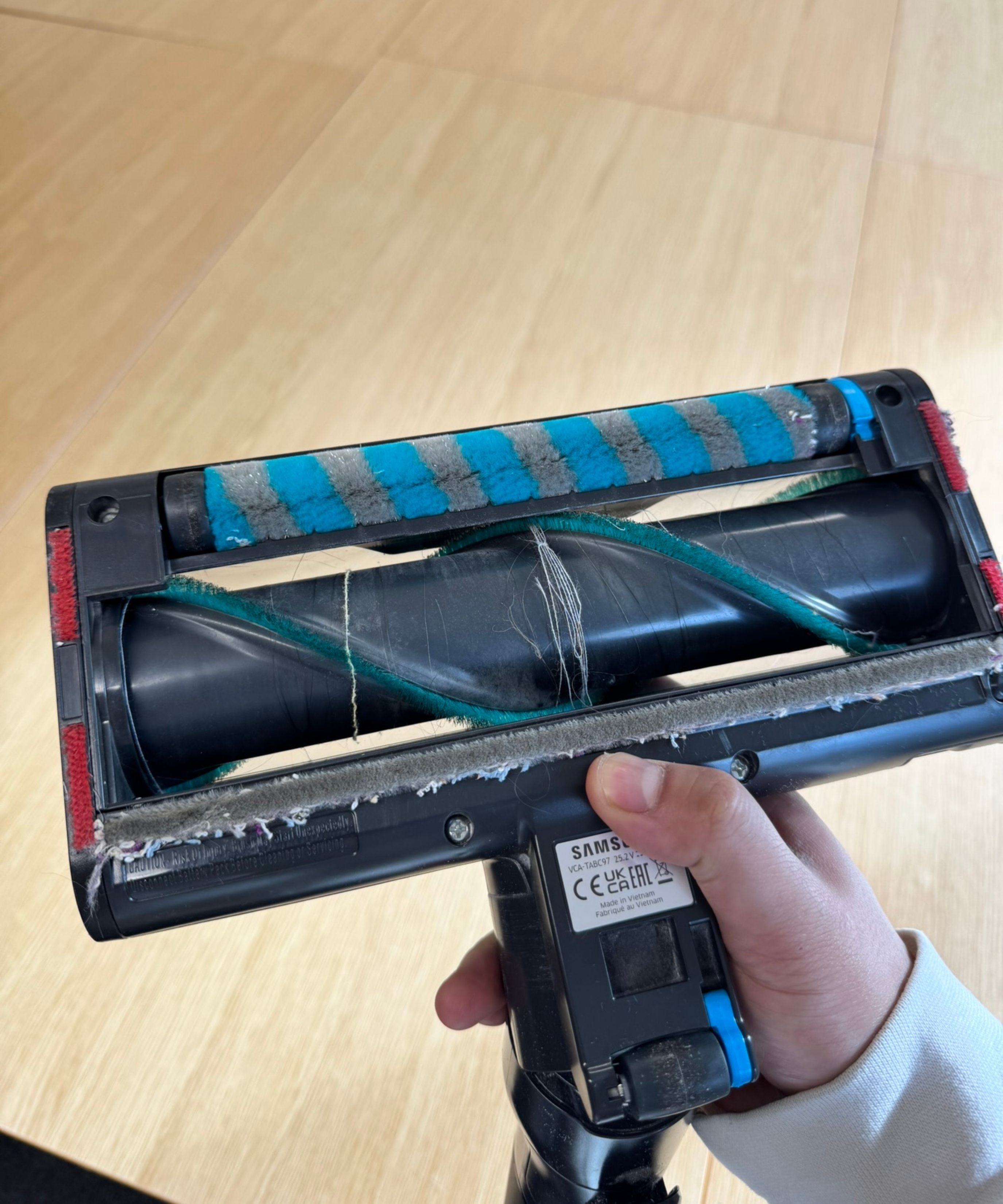 Hand holding the Samsung Bespoke AI Jet Ultra above a wooden table, showing the brushroll with thin thread and hair wrapped.
