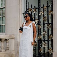 Woman wears white dress, black bag, sunglasses, and shoes. 