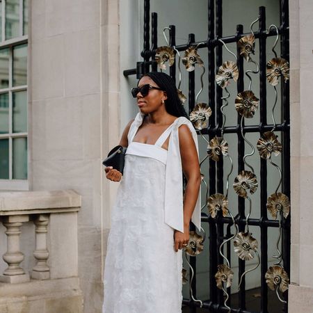 Woman wears white dress, black bag, sunglasses, and shoes. 