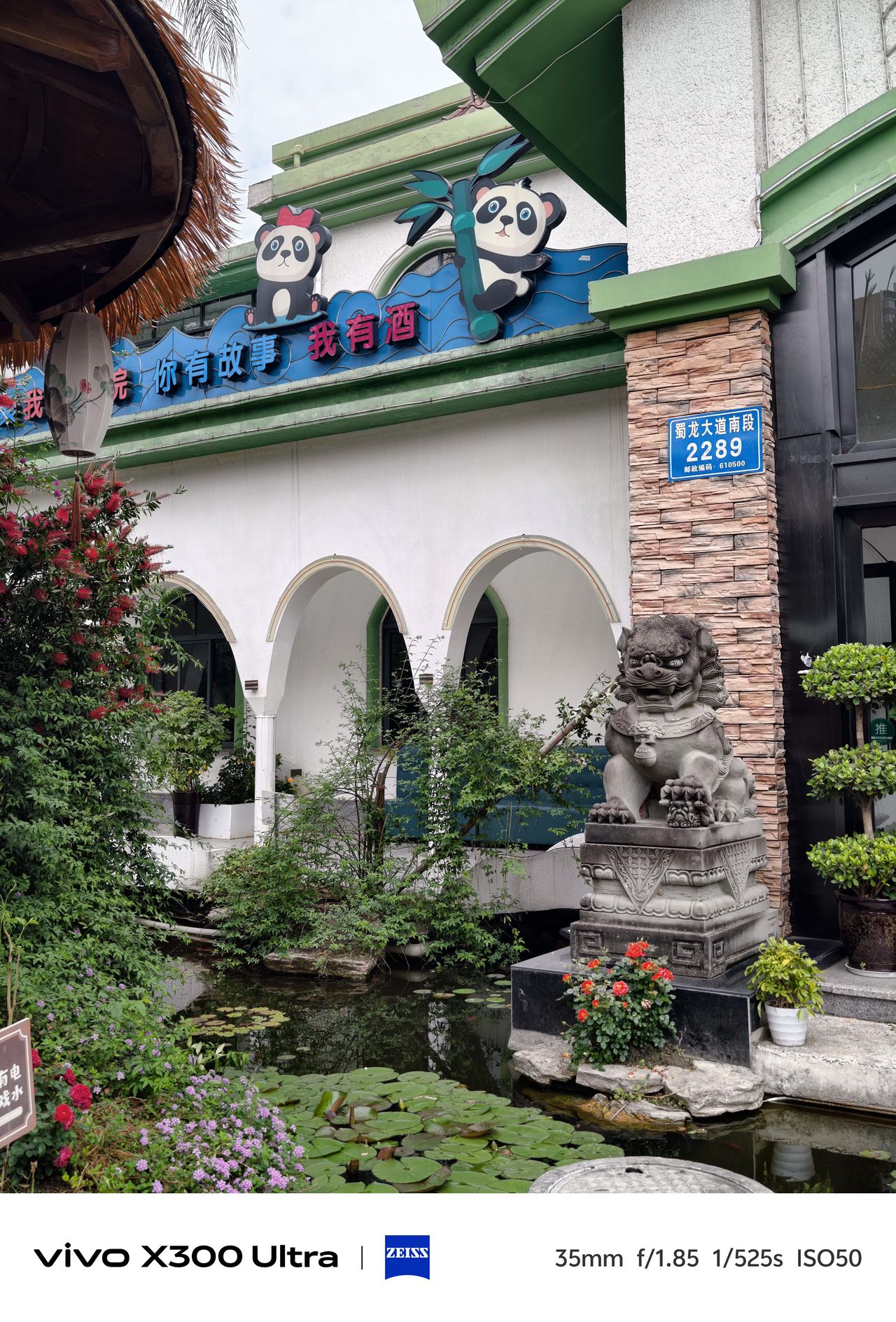 The exterior of a white building featuring arched entryways, cartoon panda decorations on the roofline, and a traditional Chinese stone guardian lion statue overlooking a small lily pond.