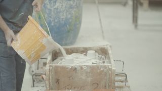 Image of an artisan hand pours liquid clay into a porous mould, yellow bucket, blurred workshop background