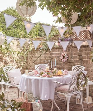 An outdoor picnic area with blue bunting, lights, a circular dining table with a white tablecloth a spread of pastel candles, eggs, and breads and cakes, and four white iron chairs with pink pillows