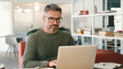 A man looks at his laptop while sitting at a desk in his office.