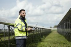 A bearded person wearing a green high-vis jacket stands in a field, holding an open laptop. Behind him, an array of solar panels sits, with a cloudy blue sky in the far distance.
