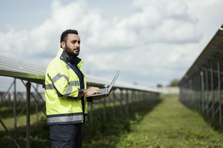 A bearded person wearing a green high-vis jacket stands in a field, holding an open laptop. Behind him, an array of solar panels sits, with a cloudy blue sky in the far distance.