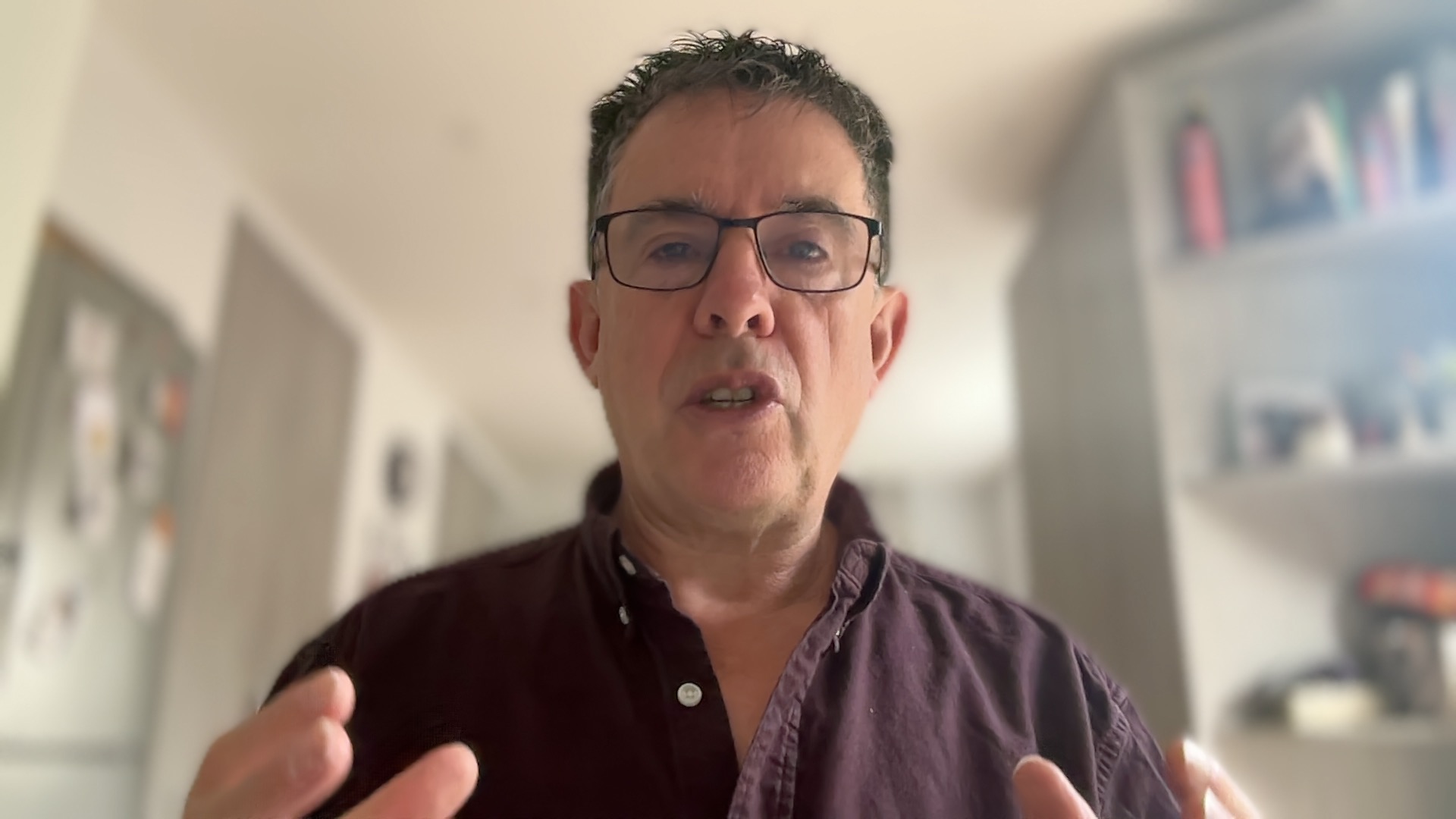 A man wearing glasses speaks to the webcam in a softly blurred home interior.