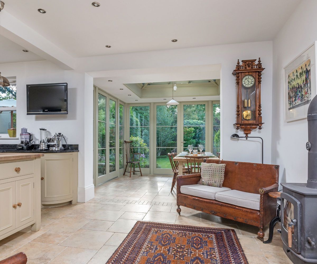 View from the kitchen through an opening into the orangery dining room beyond, with a vintage leather settle, antique wall clock, wood-burning stove and Persian rug in the foreground.