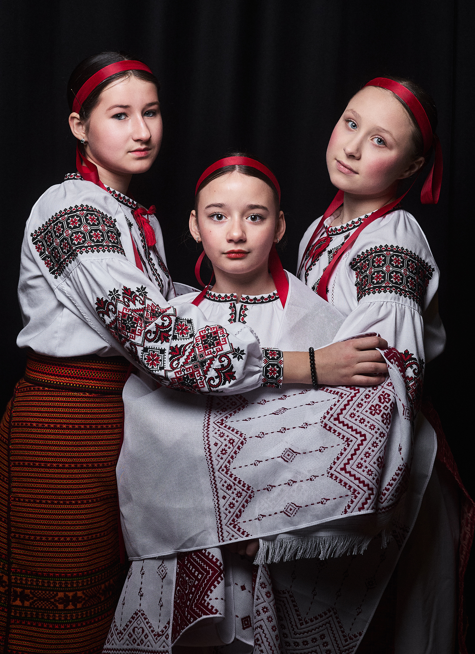 Three girls in traditional embroidered clothing and red headbands pose together against a black background