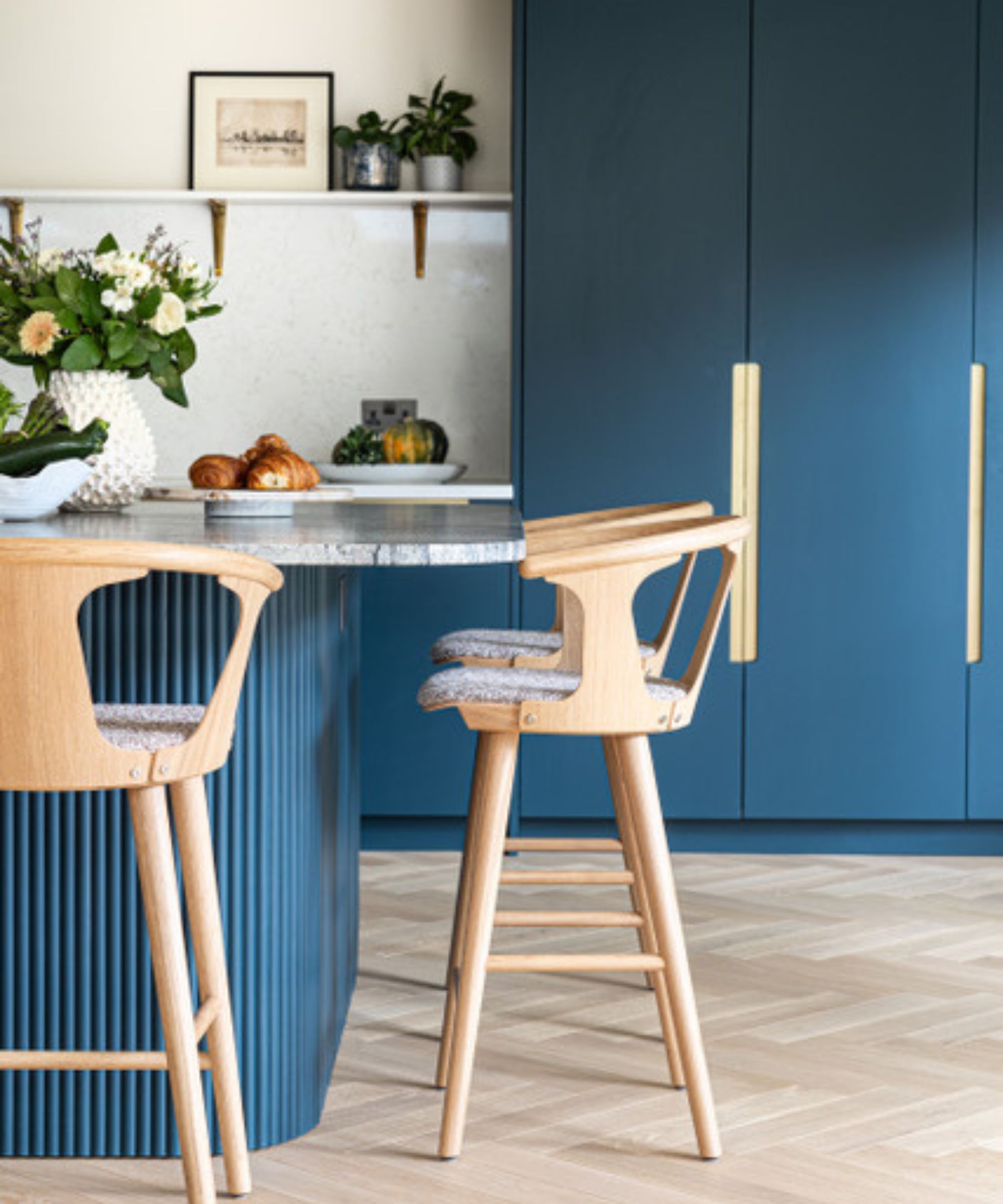 A navy blue kitchen island with fluted detailing, a stone worktop, oak bar stools and brass handles on full-height blue cabinetry with a light wood herringbone floor.