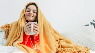 A smiling woman wrapped up in orange blanket holding white mug in both hands