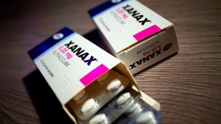 Two boxes of Xanax lying side-by-side, pointing towards the camera. They appear to be positioned on top of a wooden table. The box on the left is open and packets of pills are sliding out of it towards the camera.