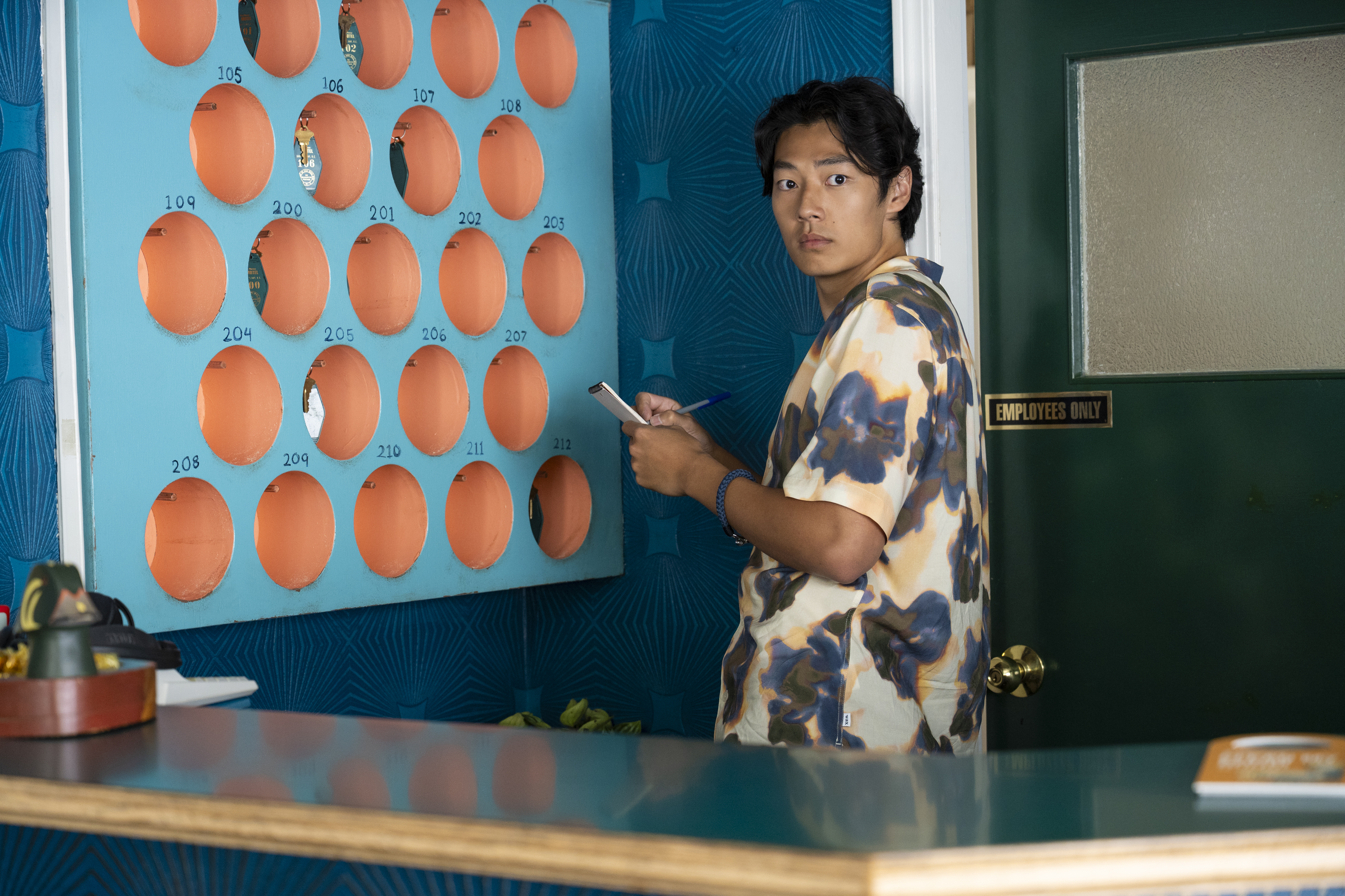 a young man in a floral printed shirt behind a motel counter writing in a notepad in a still from every year after