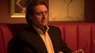 Ian McShane's Winston sits at a table looking at someone off-camera in John Wick