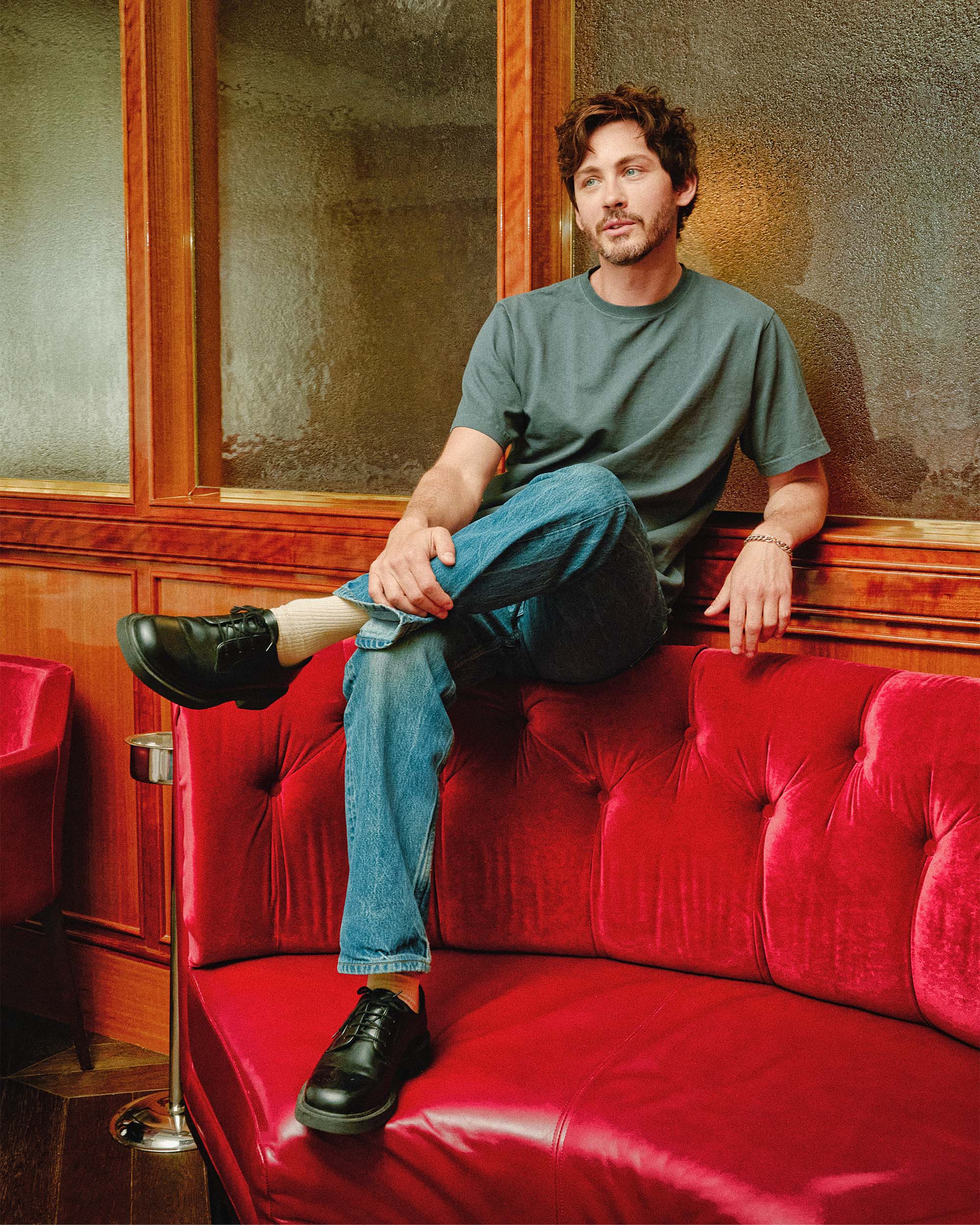 Logan Lerman sits cross legged on top of a red velvet booth