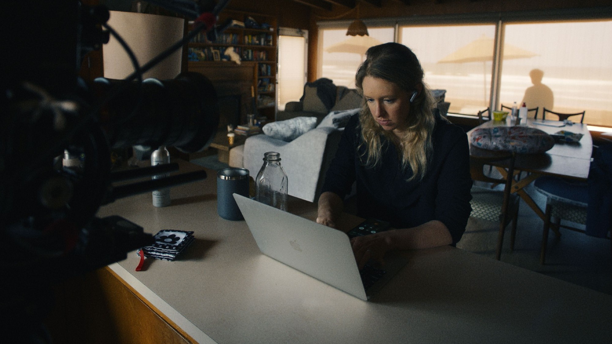 Elizabeth Holmes sitting at a laptop, with a large camera focused on her with Nathan Fielder in the back, looking through a window in You Can See Everything