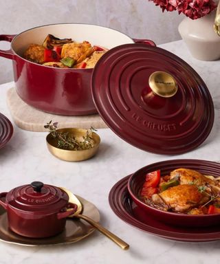 A chicken pot roast in a burgundy Le Creuset Dutch Oven; chicken served on burgundy plates; a mini burgundy cocotte; all on a white marble countertop.