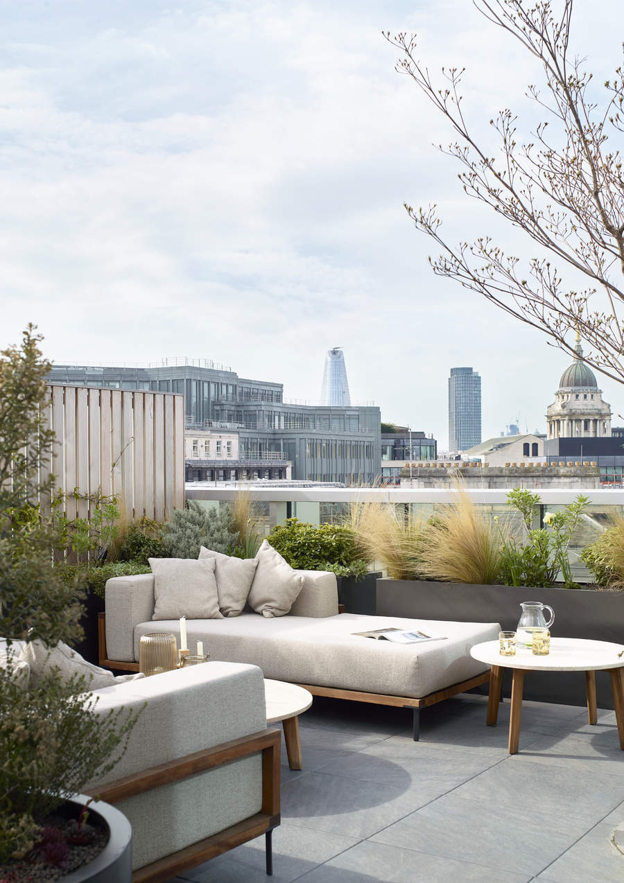 Image of a sunny balcony with wooden lounge furniture that has thick, beige cushioning. There are two round, white and wooden coffee tables and lots of plants.