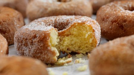 Half-eaten sugar-coated doughnut is shown in the center of the image. It is on what appears to be a sheet on baking paper with other untouched donuts surrounding it. The other donuts are slightly blurred. 