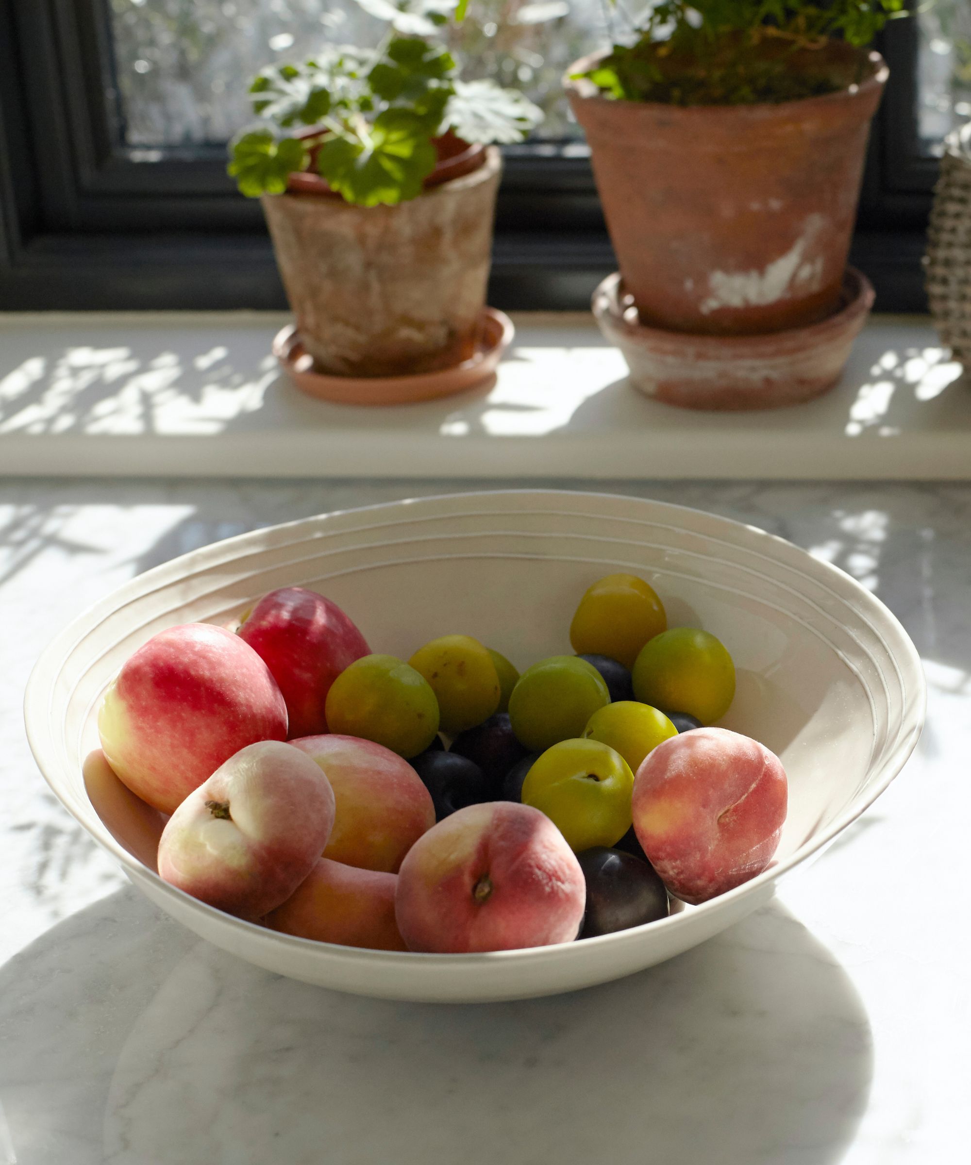White stone fruit bowl on white marble surface, filled with peaches and plums