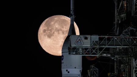 The tip of NASA's Space Launch System rocket is visible with the full moon rising behind it in the black night sky. The steel framework of the launch tower dominates the right of the screen, and the crew arm is extended, bridging the gap to the rocket's white upper stage.