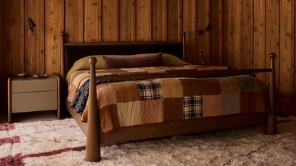 Image of a wooden bed with a patchwork quilt on it in a room that has wooden paneled walls. There is a white and red shag rug on the ground.