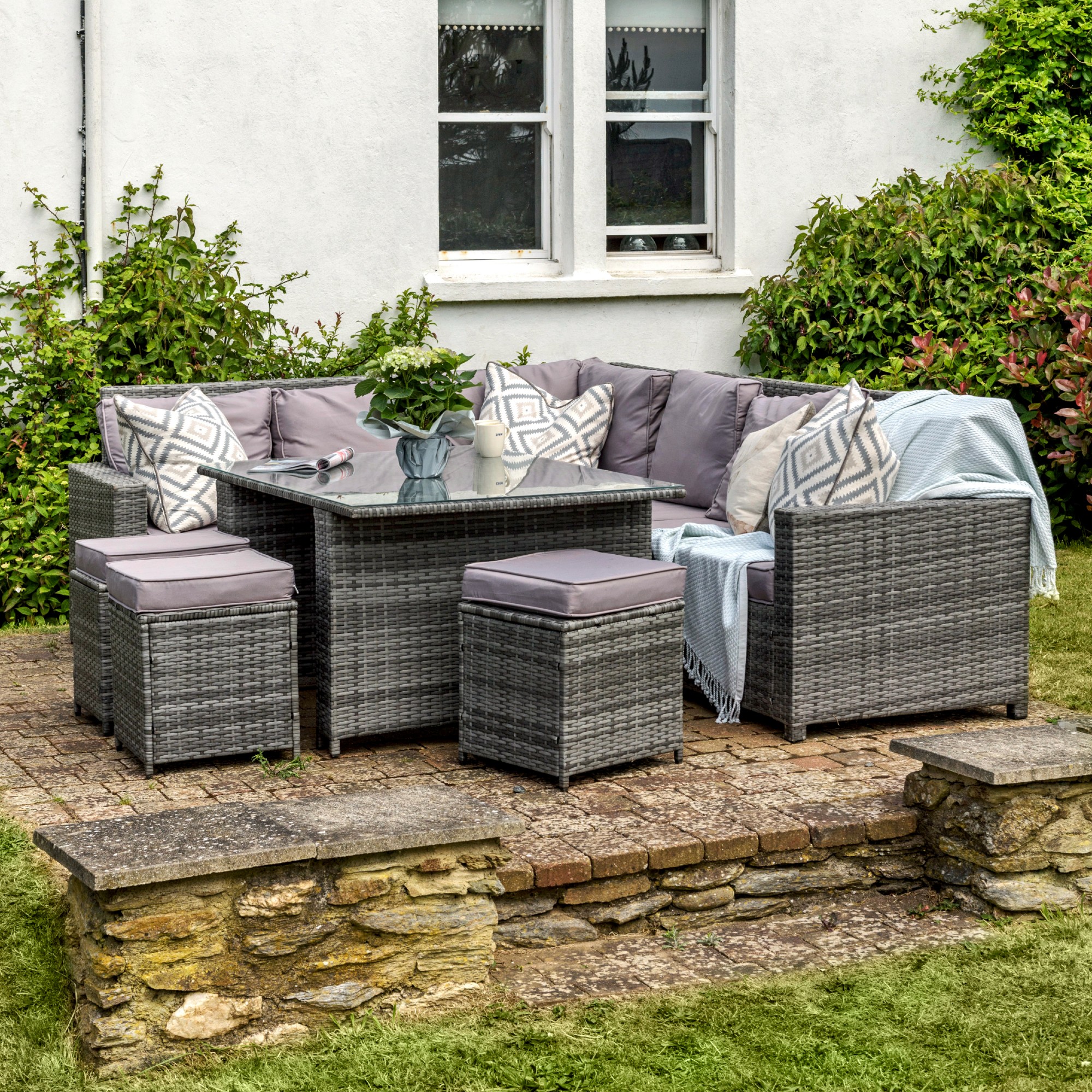 A garden furniture set in grey rattan effect of a corner sofa, a table and three stools with grey seat and back cushions and patterned scatter outdoor cushions