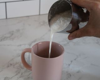 Contributing editor, Camryn Rabideau, a caucasian woman pouring frothed milk from a stainless steel metal pitcher into a pastel pink ceramic mug which is placed on a marble effect kitchen countertop