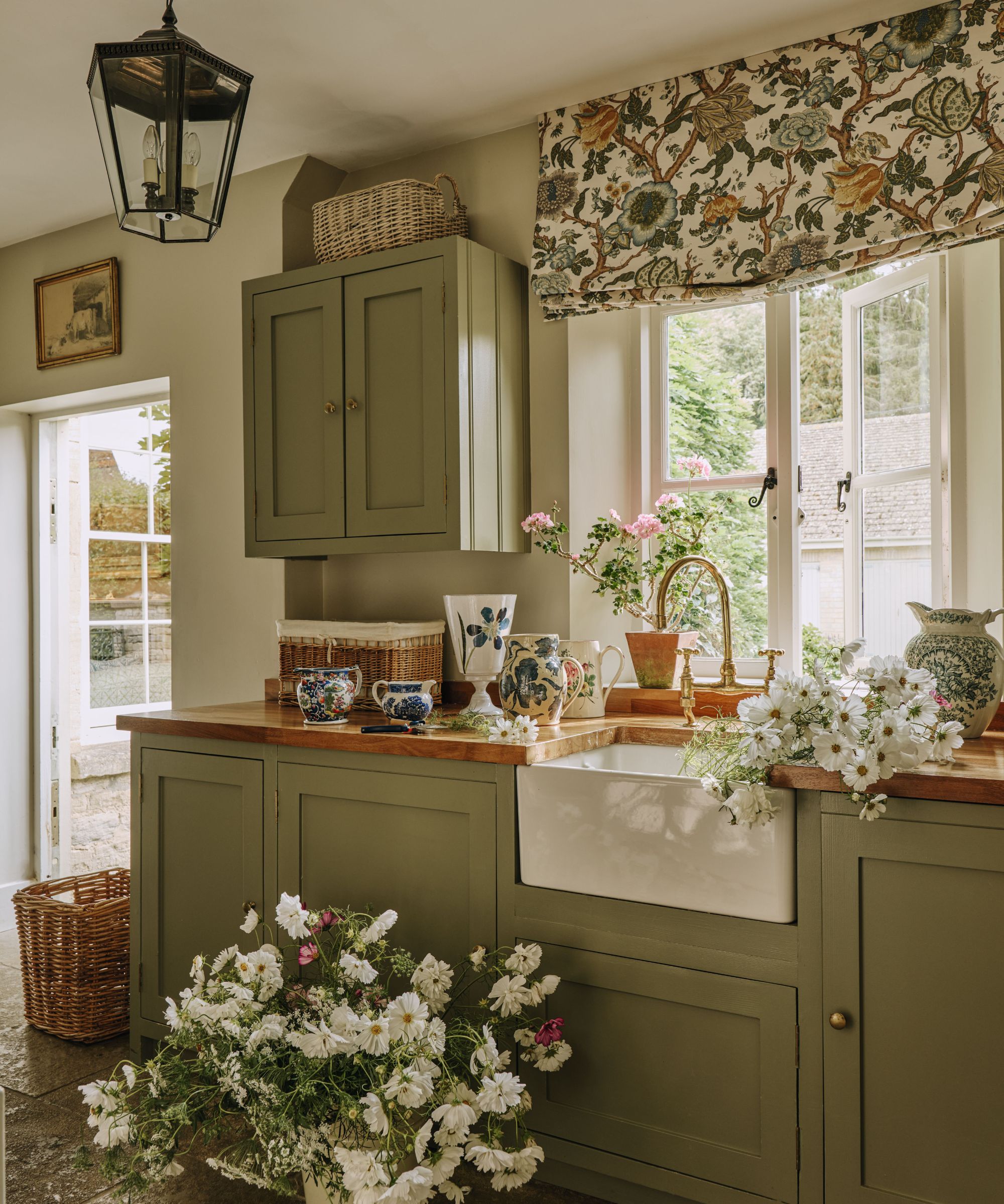 Rustic kitchen with sage green cabinets, a apron-front sink, wooden countertops, floral window valance, and fresh white flowers