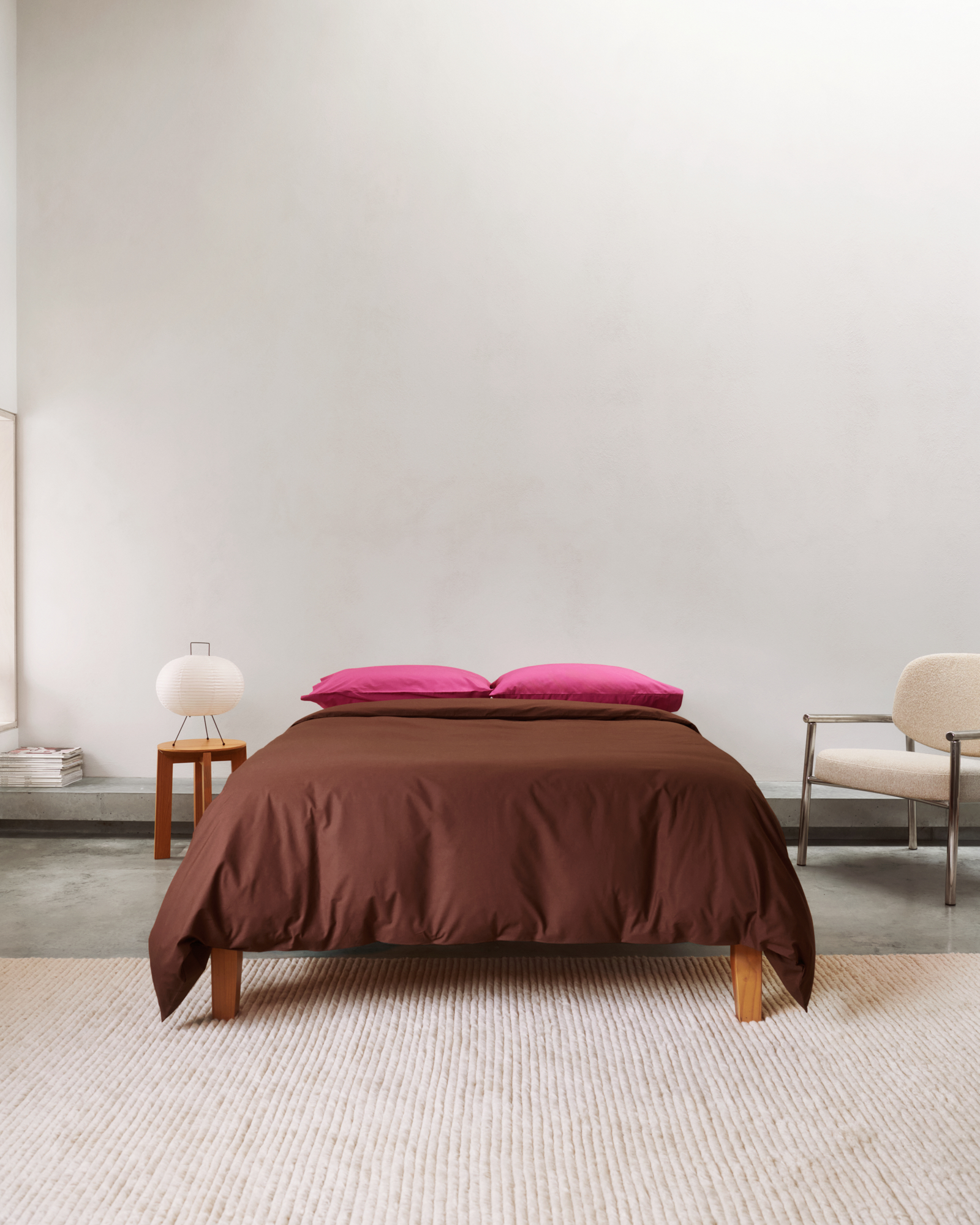 image of a white bedroom with a bed in the middle that has brown bedding and hot pink pillows. There is a white chair to one side of the bed and a wooden side table with a lantern to the other.
