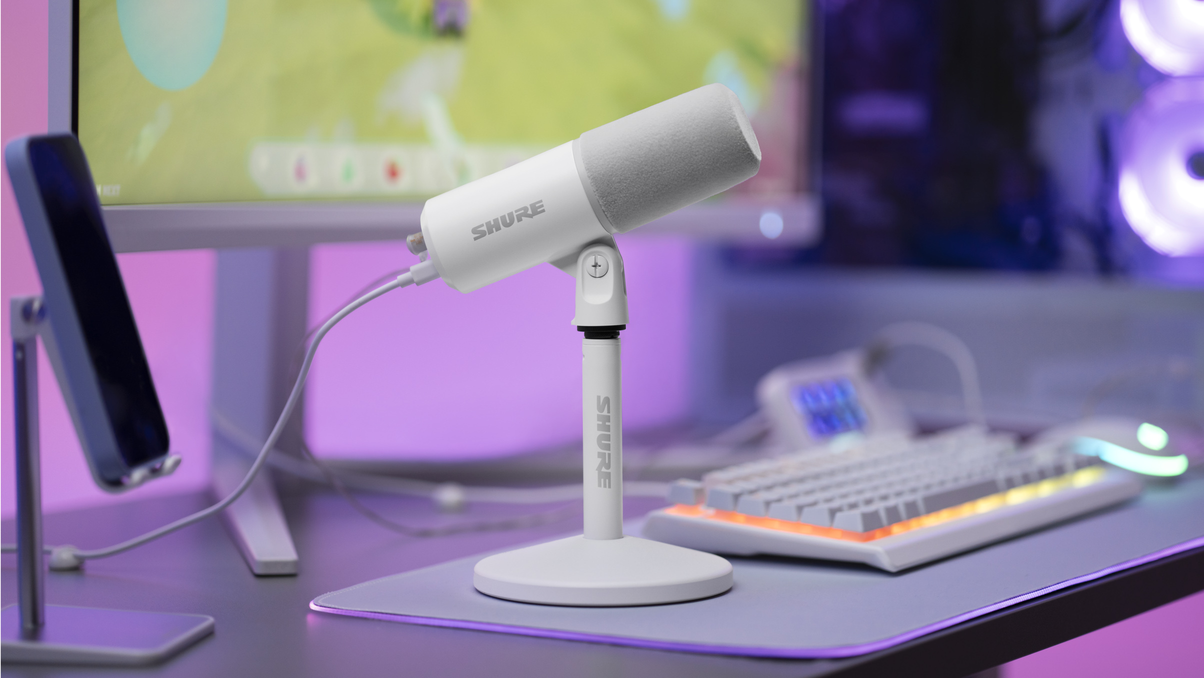 White Shure MV6 USB Gaming Microphone Gen 2 on a desktop stand at a gaming setup with a keyboard, monitor, and purple ambient lighting.